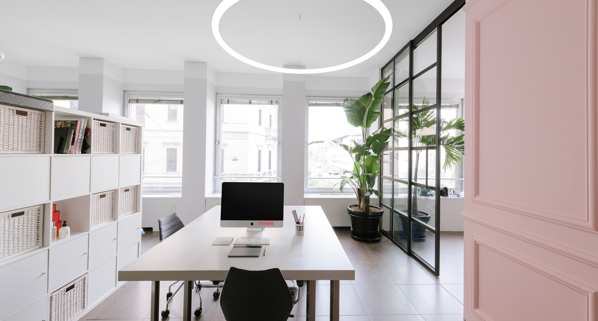 Modern office with Artemide circular LED pendant light illuminating a white desk, shelving, and green plants by large windows.