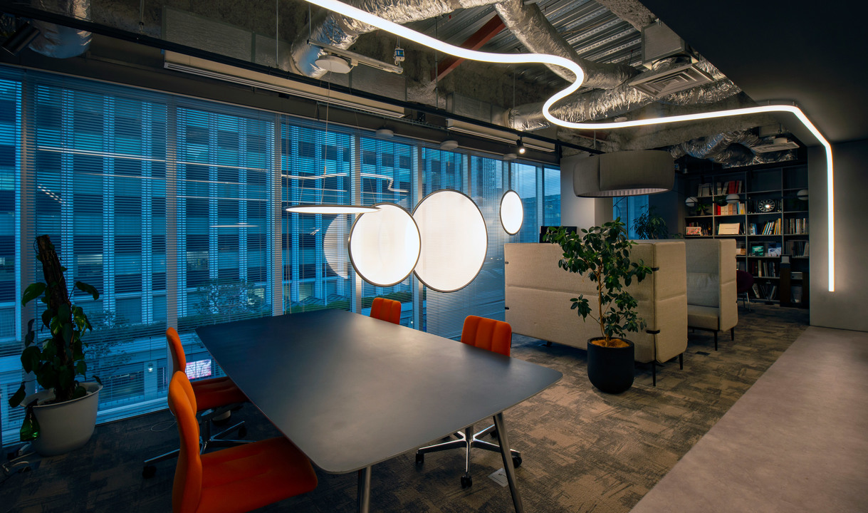 Modern office meeting room with Artemide circular pendant lights and sinuous LED ceiling strip over a dark conference table with orange chairs