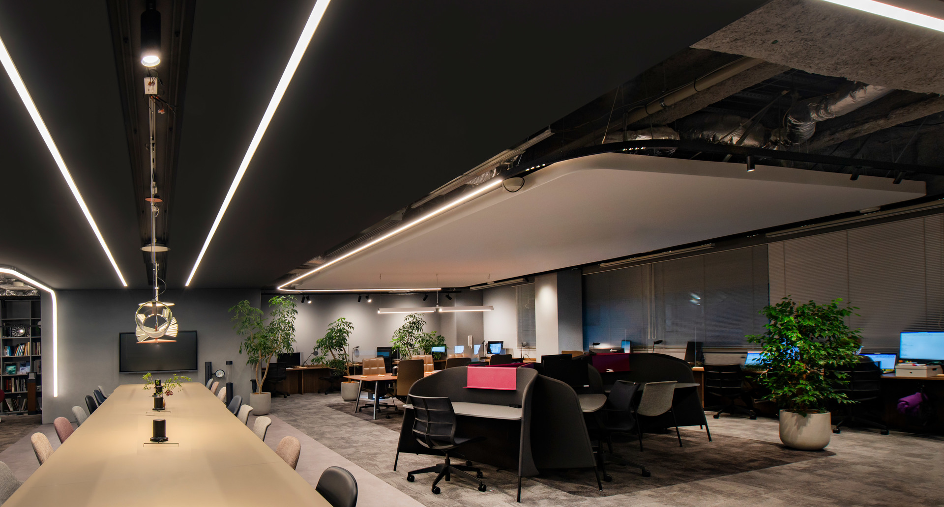 Modern office with dark ceiling featuring linear LED light strips illuminating a long conference table and open workstations with plants.