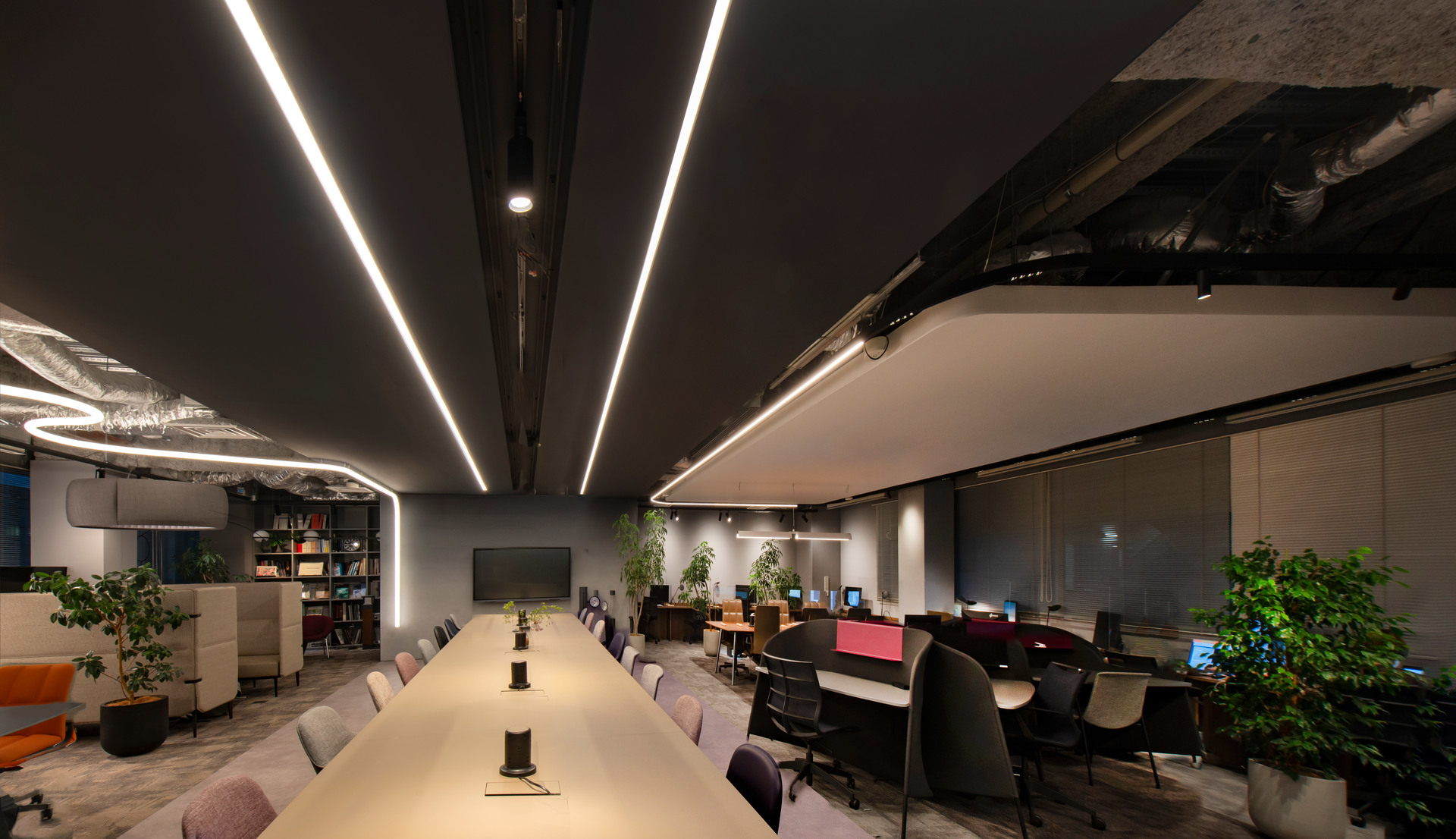 Modern office with long conference table lit by linear LED ceiling lights, workstations, plants, and dark exposed ceiling.