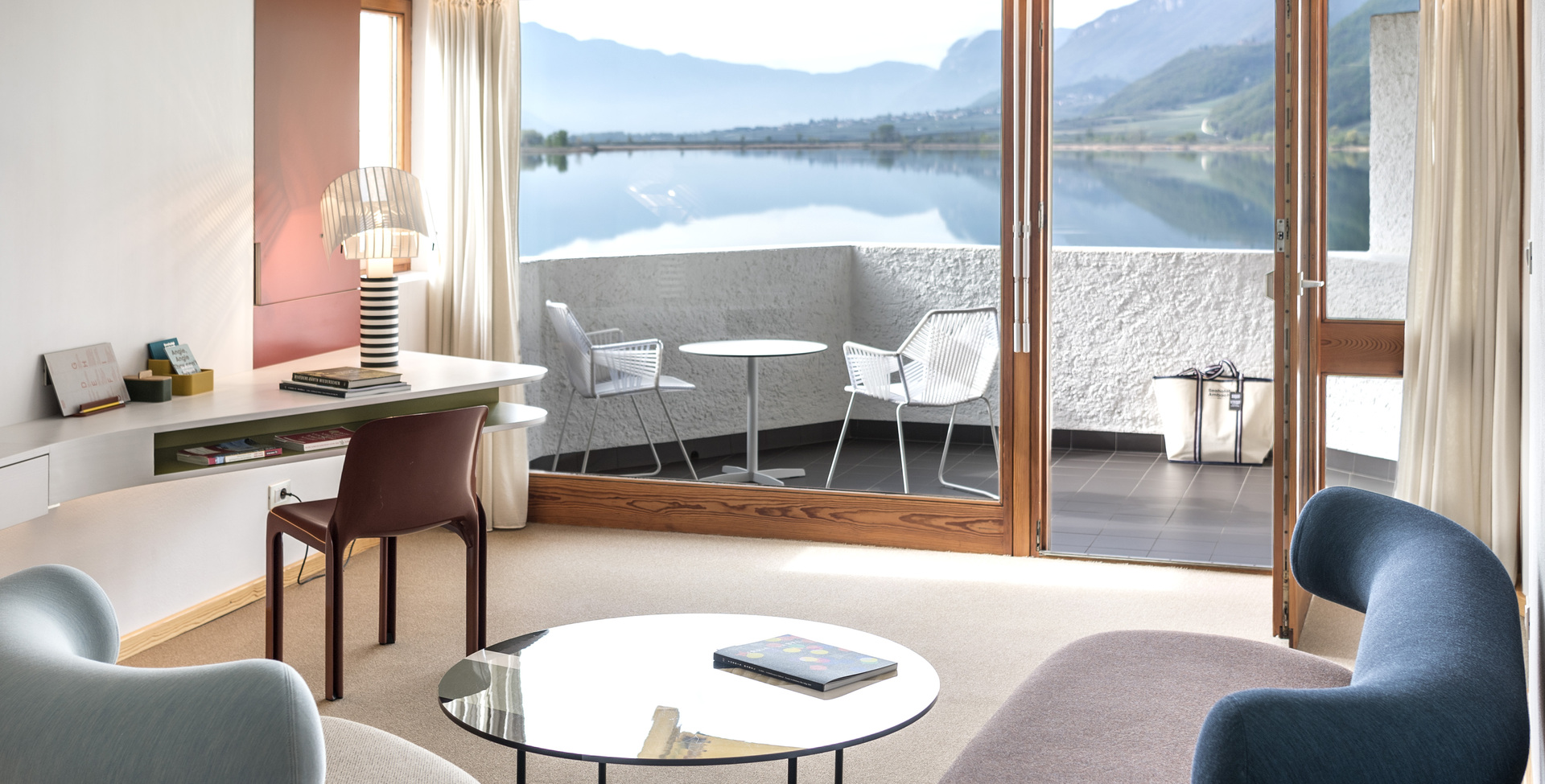 Modern hotel suite with Artemide table lamp on desk, blue armchairs, glass coffee table, and panoramic lake and mountain view through floor-to-ceiling windows