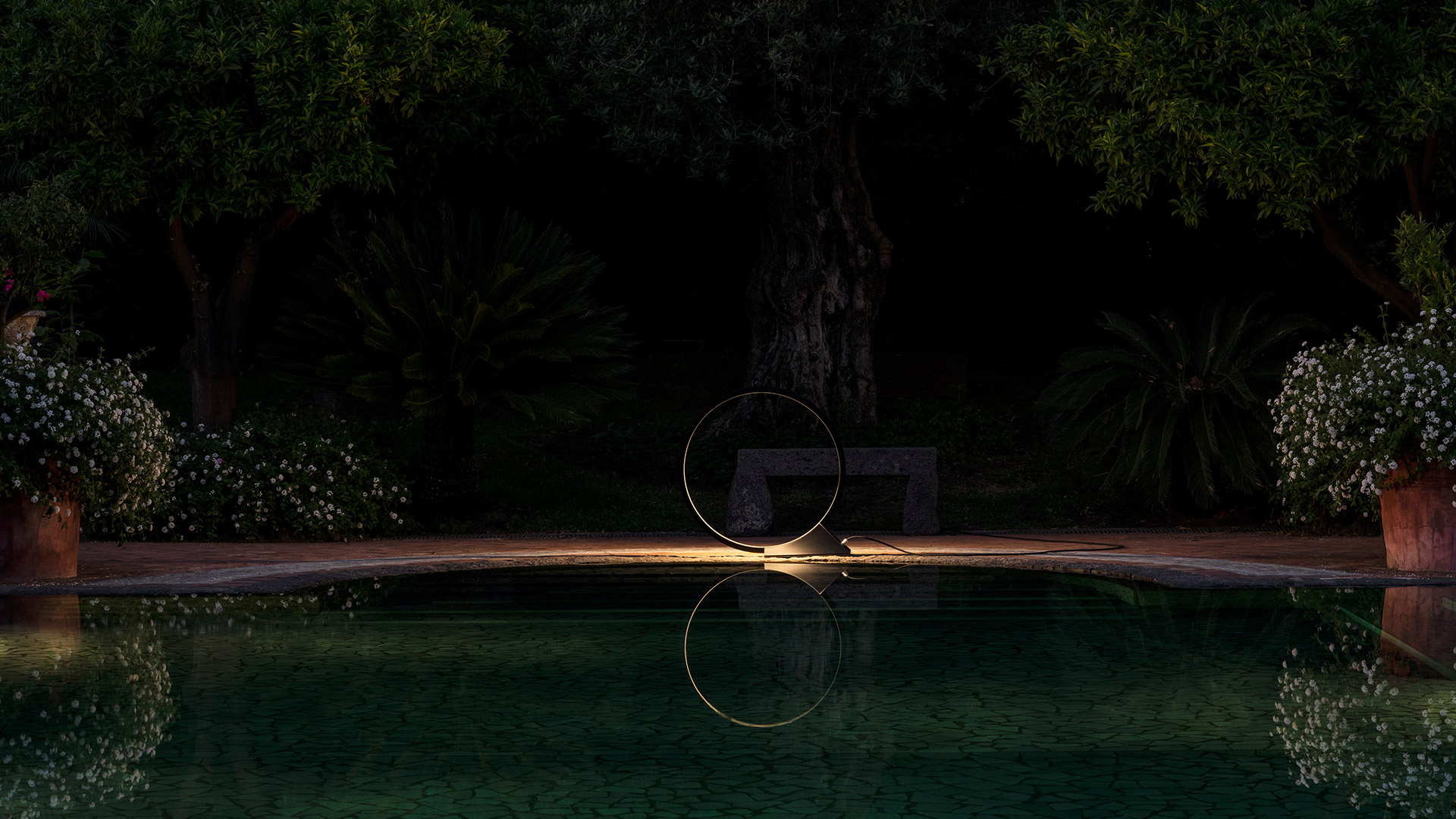 Artemide 'O' outdoor floor lamp: a large black circular ring emitting warm light, standing on grass between trees at dusk