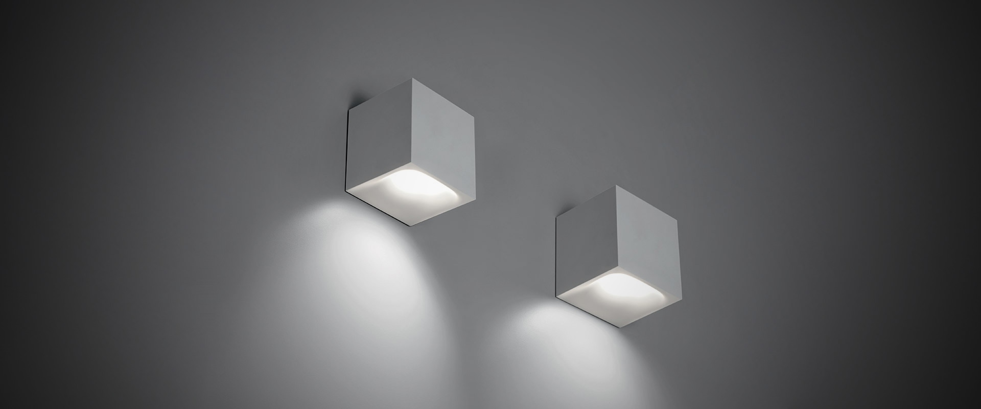 Two Artemide Aede cubic wall lights in white, casting downward beams on a dark grey wall