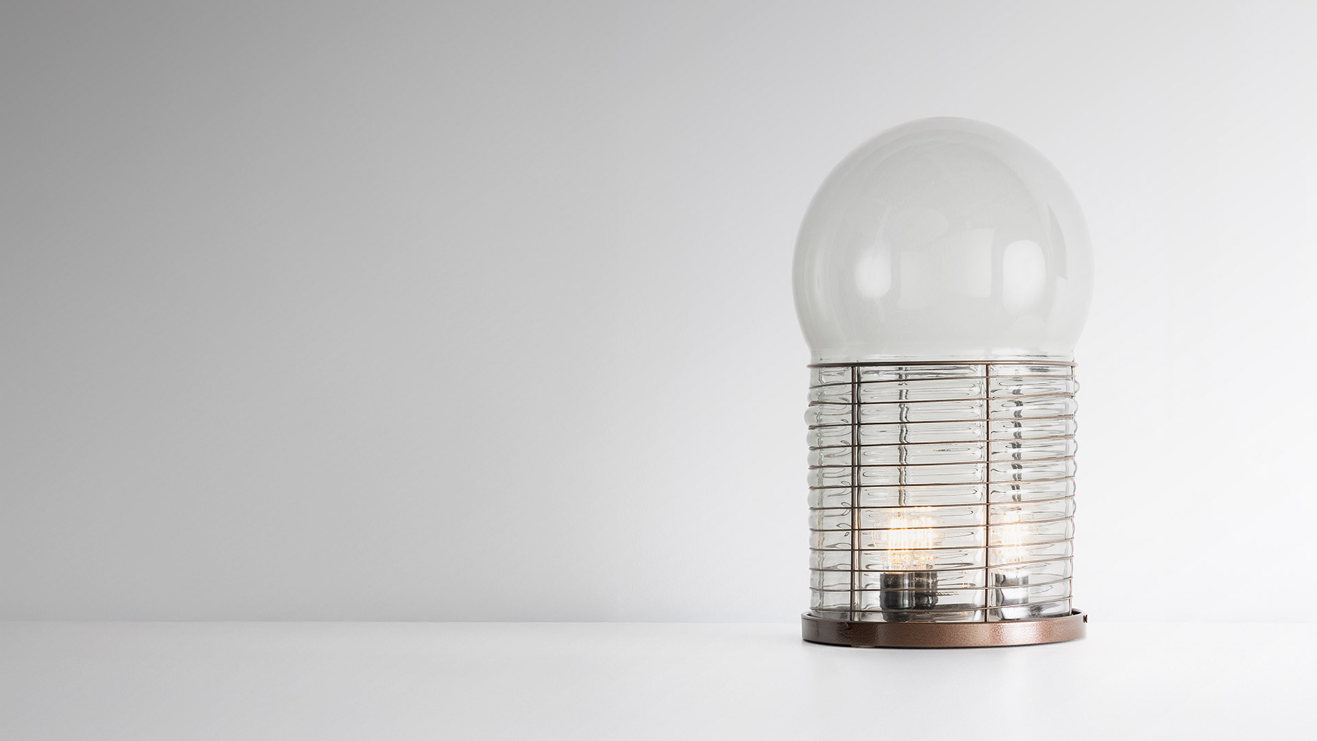 Artemide Alcinoo table lamp: clear glass sphere atop a cylindrical ribbed glass base with visible bulbs, emitting soft blue ambient light