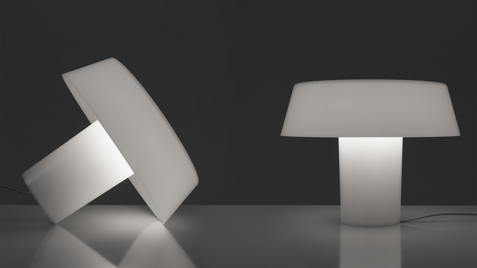 Two Artemide Amami table lamps: left tilted disc-shaped lamp, right upright mushroom-shaped lamp, both in white diffused light