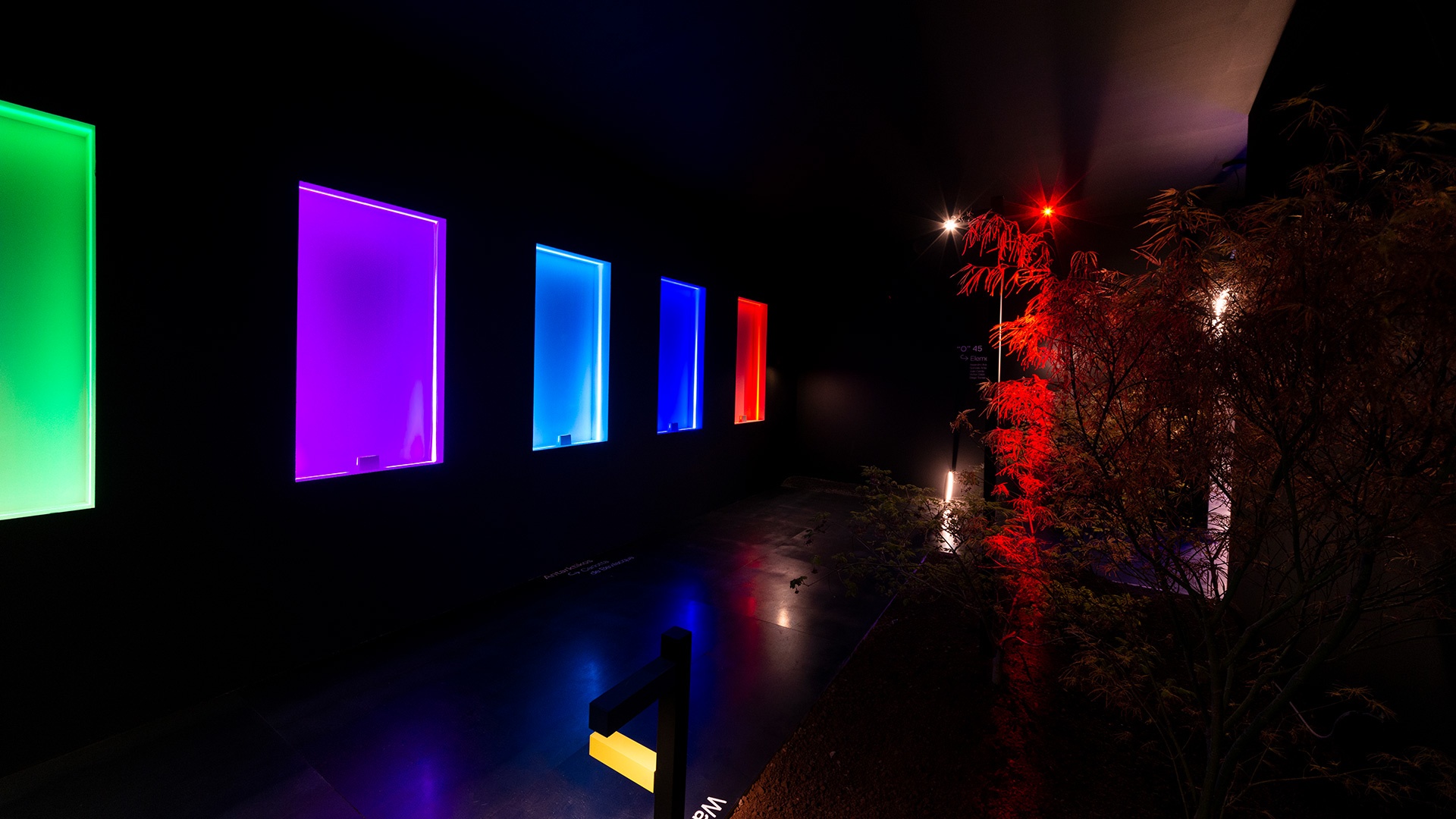 Artemide Antarktikós outdoor installation: colorful illuminated rectangular panels in green, purple, blue, and red against a dark background with lit foliage