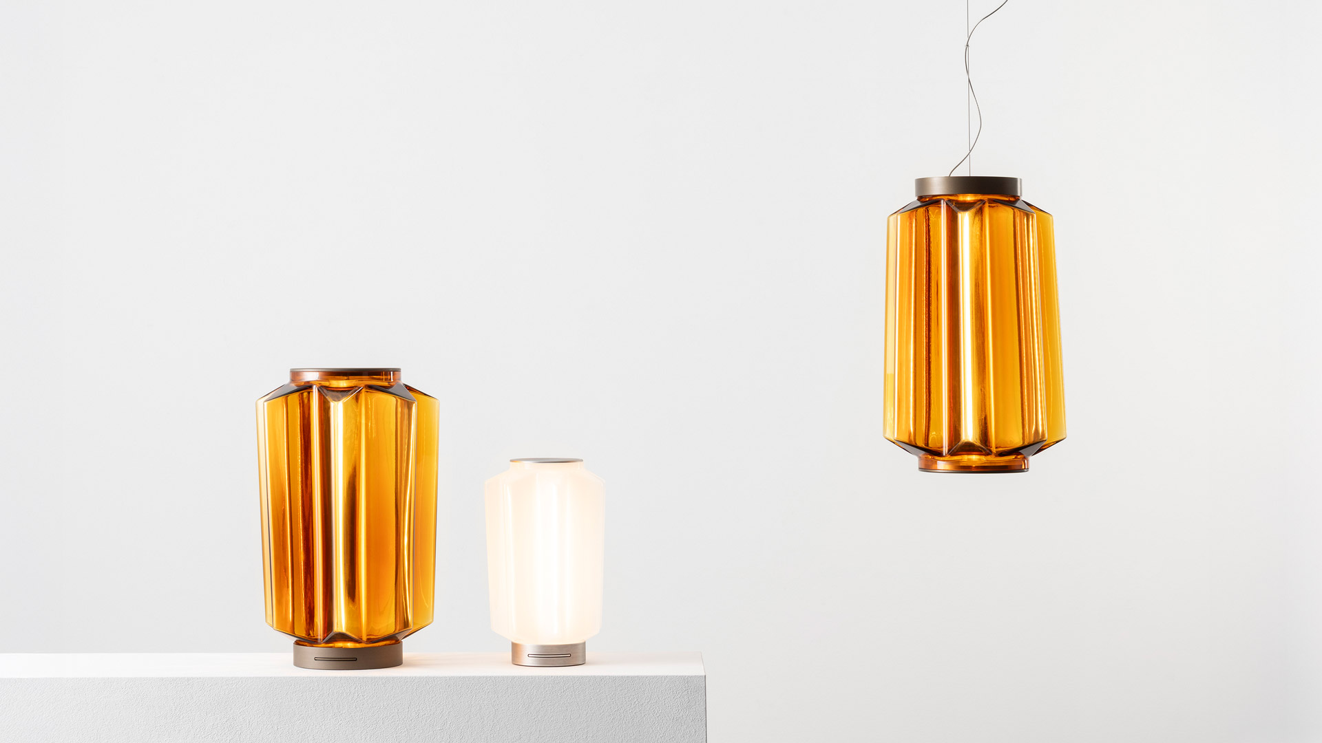 Artemide Auralia table lamp in amber glass with geometric faceted body, glowing warmly on a wooden table beside a small wooden sculpture