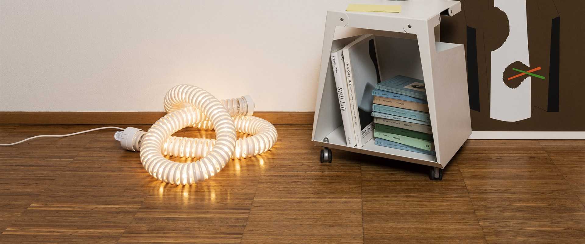 Artemide Boalum lamp: flexible luminous white tube coiled on wooden floor, emitting warm light, next to a white storage unit