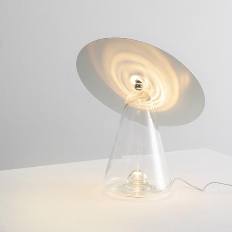 Artemide Boltons table lamp: clear glass cone base with chrome disc shade tilted upward, casting spiral light pattern on surface