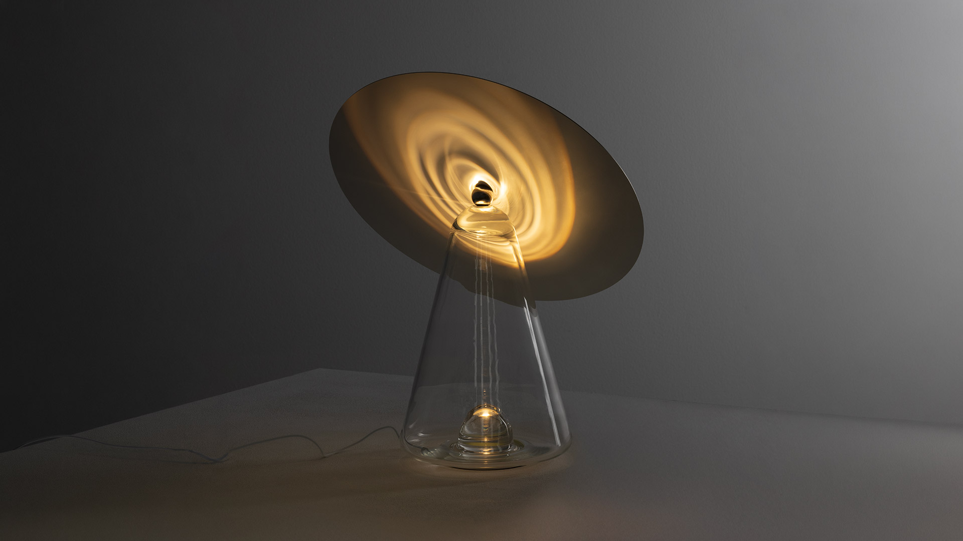 Artemide Boltons table lamp: tilted golden disc reflector over a transparent conical glass base with warm glowing light source