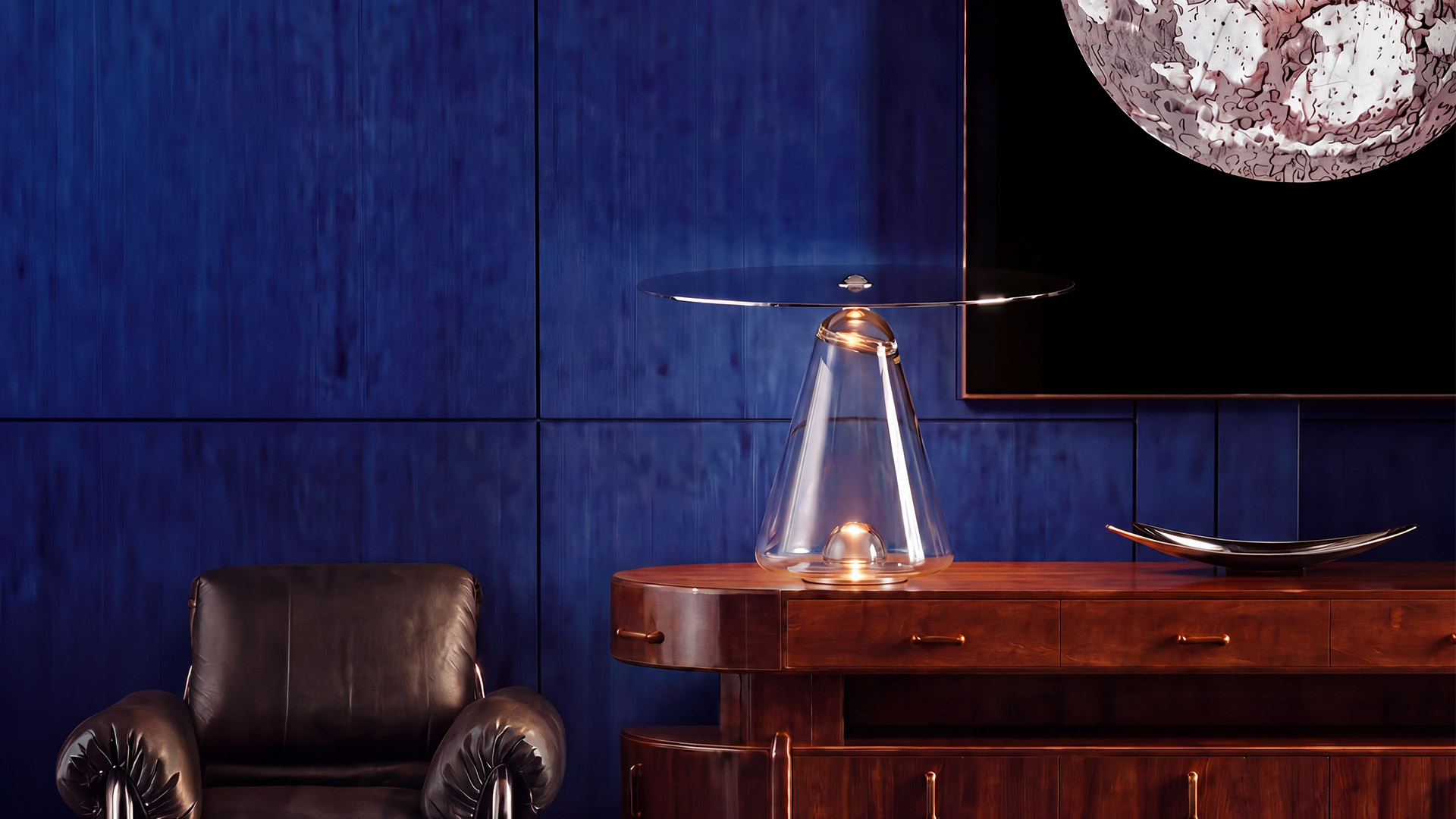 Artemide Boltons table lamp: transparent conical glass base with warm bulb glow, topped by a tilted large circular grey metal disc shade