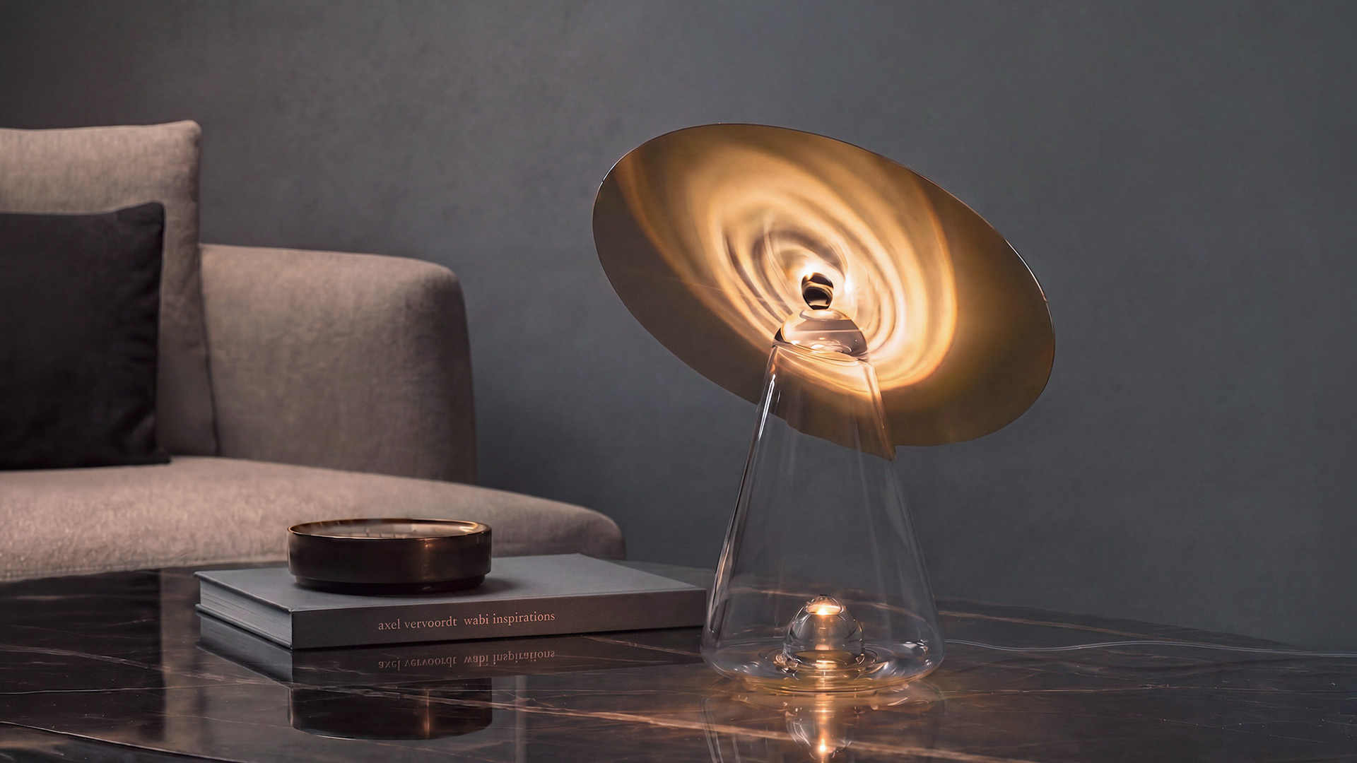 Artemide table lamp with clear conical glass base and tilted white disc shade, glowing on a marble coffee table in a modern living room.