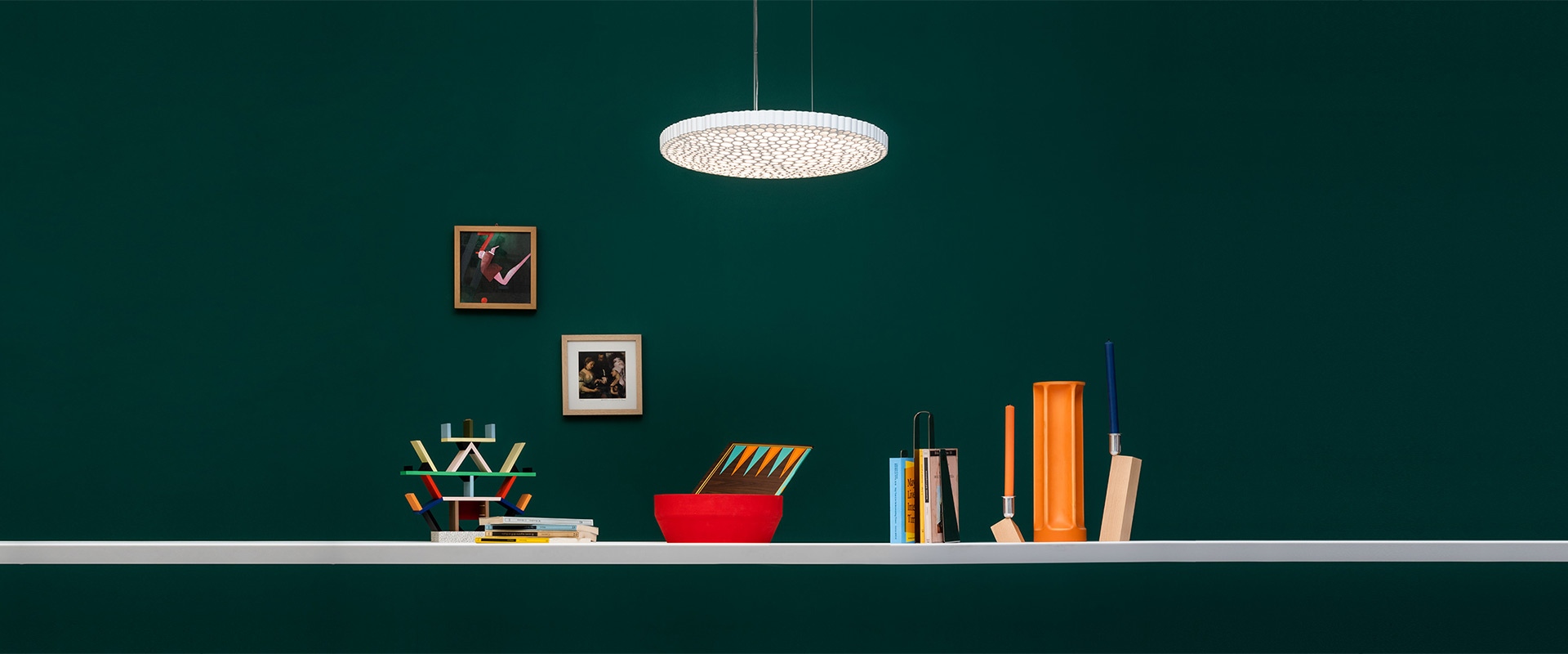 Artemide Calipso pendant lamp with white perforated disc shade hanging above a shelf with books, colorful objects, and framed artwork on a dark green wall