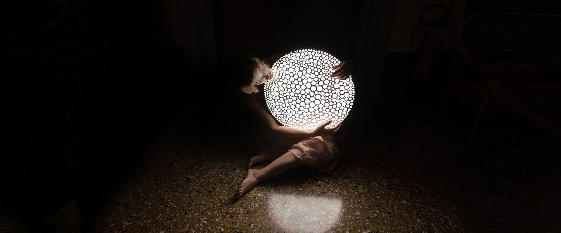 Person cradling a glowing Artemide Calipso spherical lamp with organic perforated pattern, casting light on a dark floor