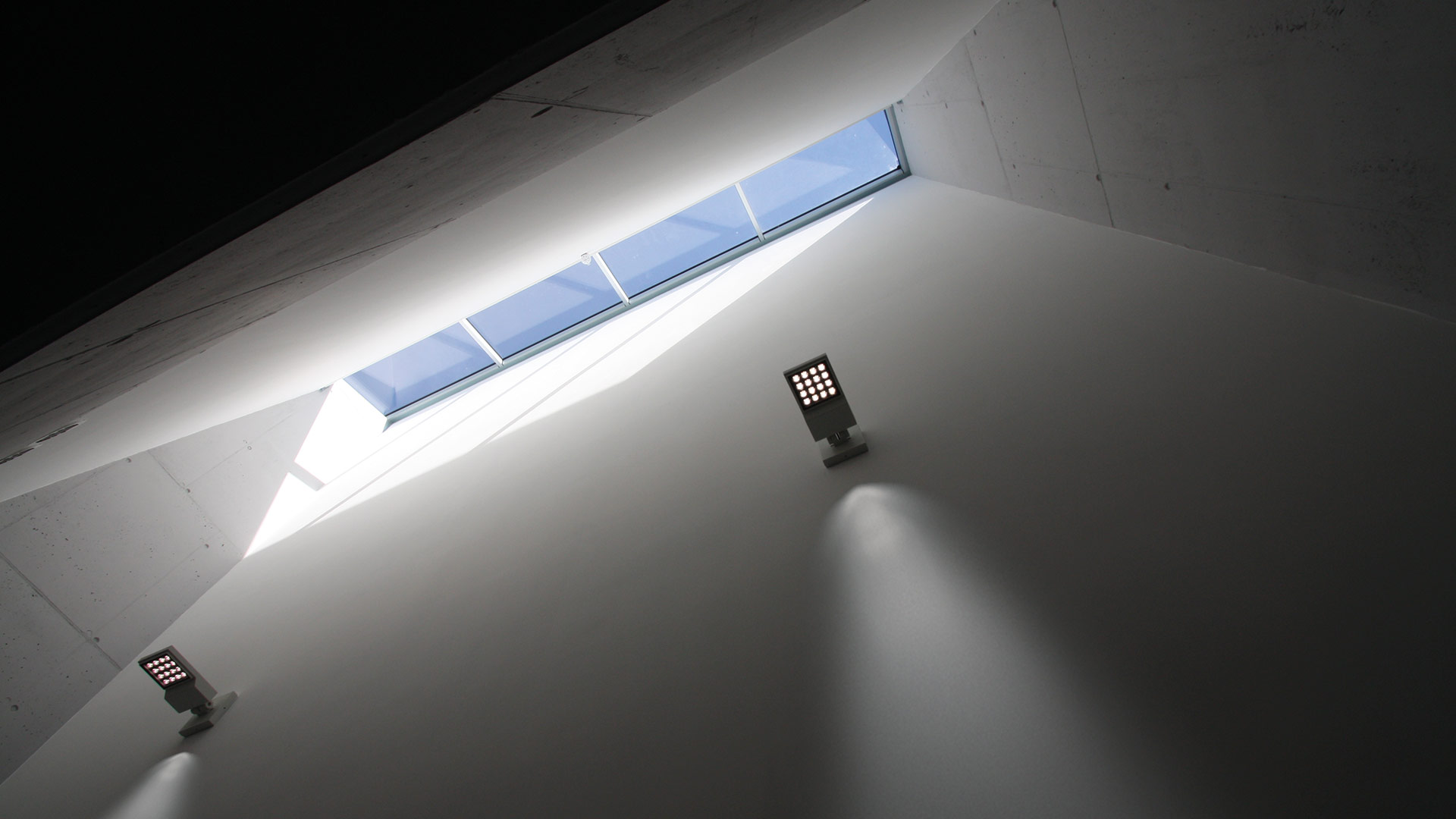 Two Artemide Cefiso wall-mounted LED spotlights illuminate a white angular wall beneath a narrow skylight with natural light.