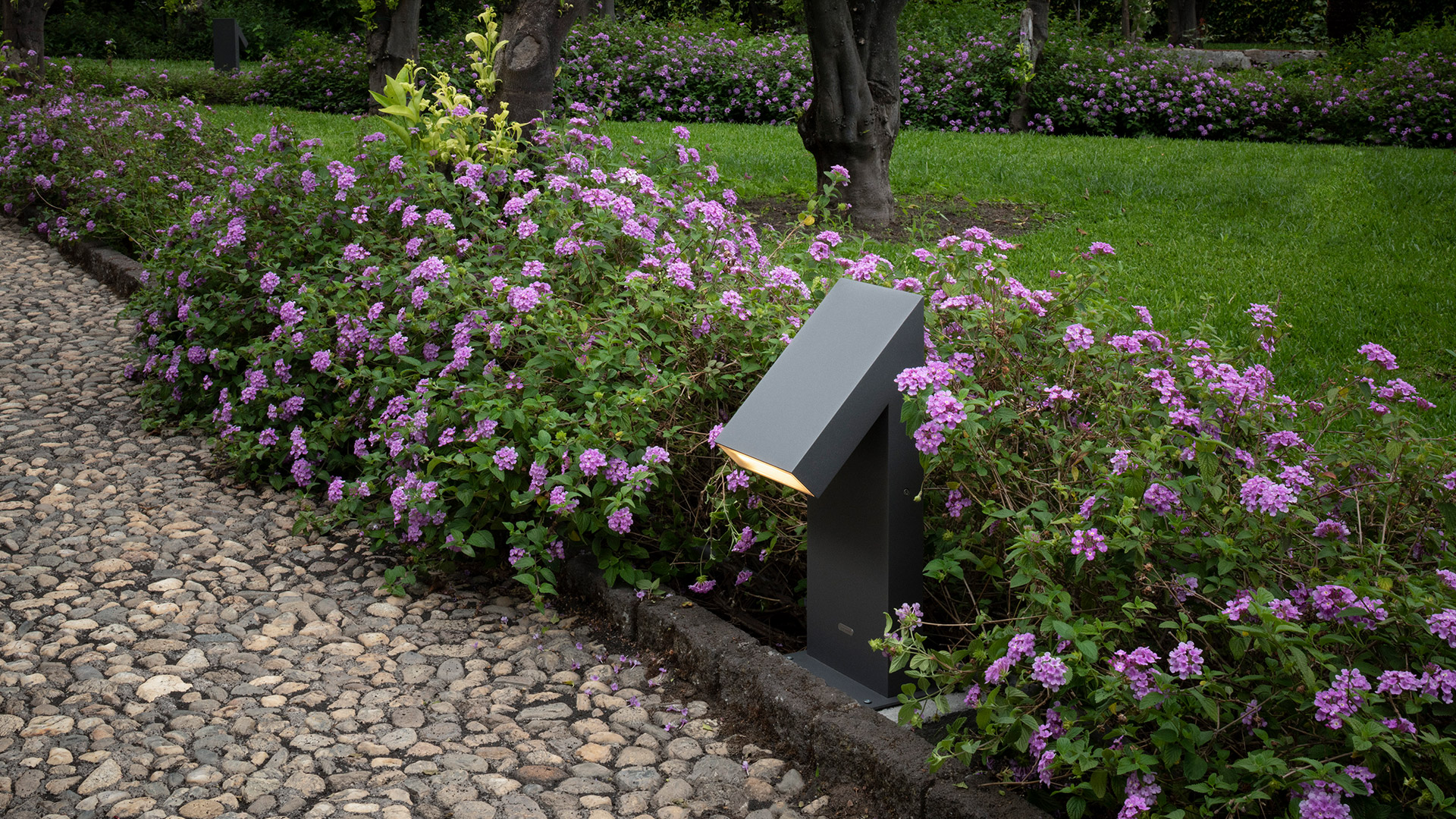 Artemide Chilone bollard lights illuminate a lakeside terrace at dusk, with benches, cobblestone path, and mountain panorama in background
