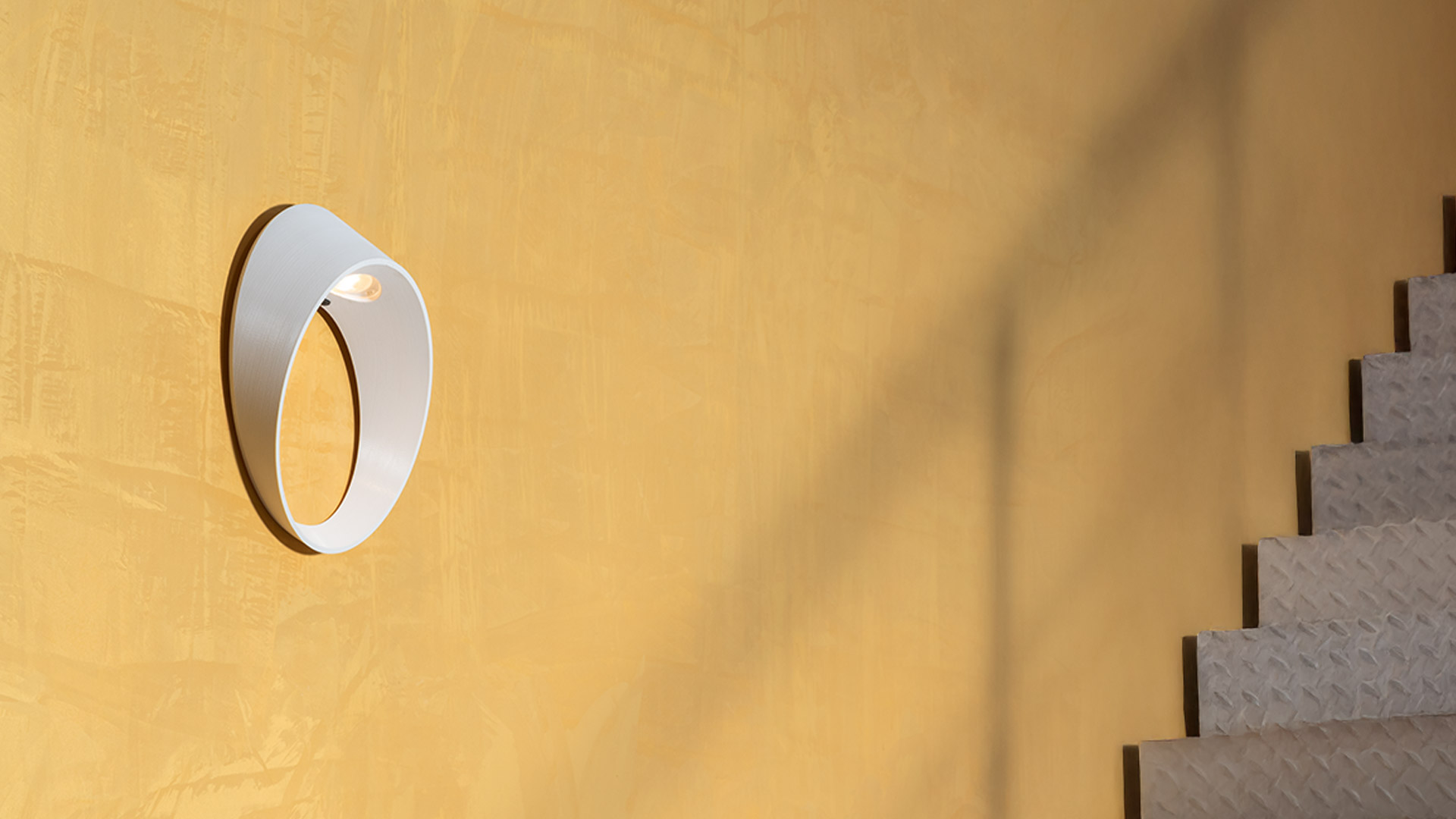 Artemide Cleomede wall lamp, circular taupe ring-shaped diffuser emitting warm light, mounted on dark wall with plant