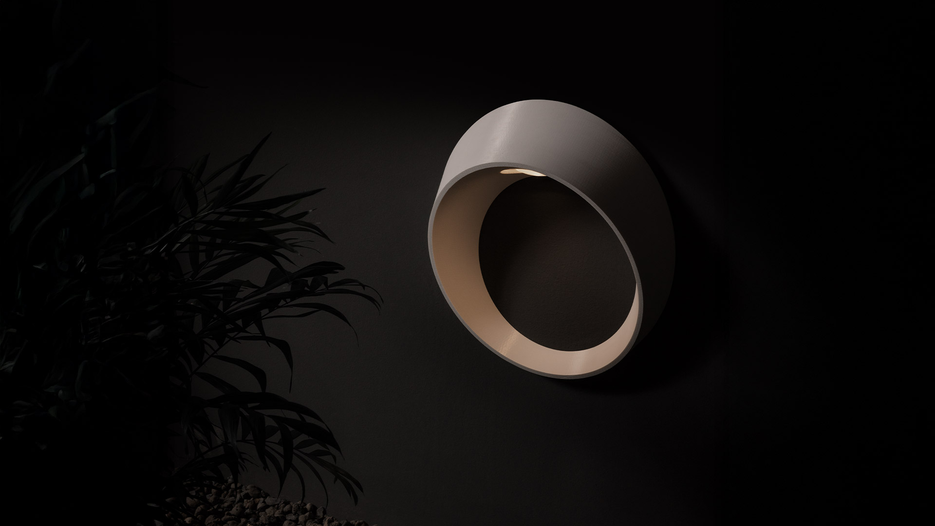 Artemide Cleomede wall lamp: circular dark bronze fixture emitting warm downward light from a small aperture, mounted on dark wall
