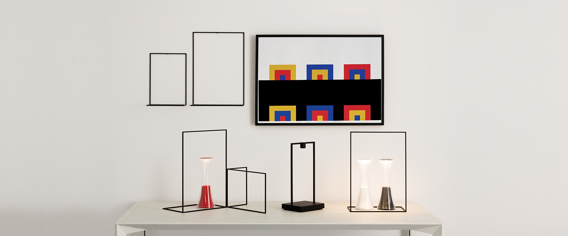 Artemide Curiosity lamp: black rectangular frame with integrated LED base light, illuminating a plant, ceramic bowls, and a glass pitcher on a white surface
