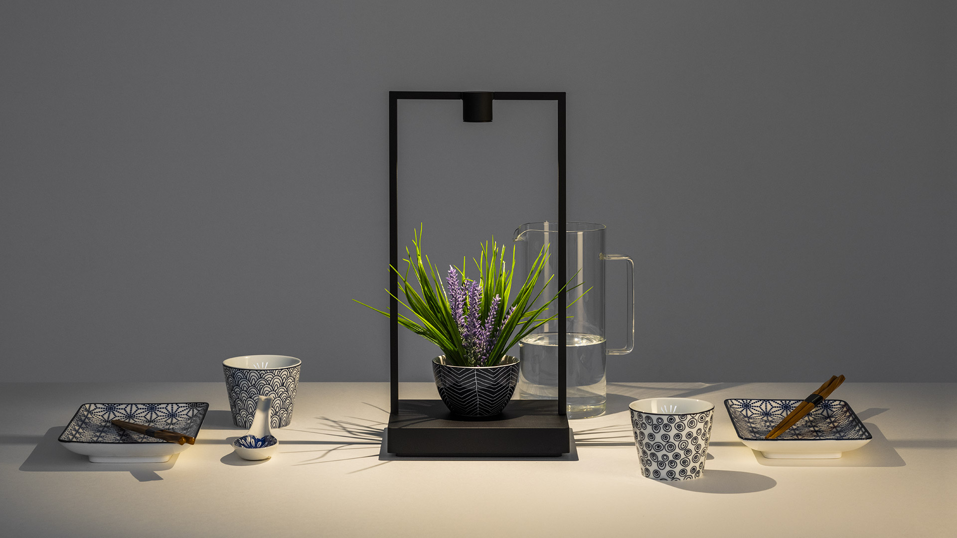 Artemide Curiosity black table lamp with rectangular frame on dark base, displayed on a white side table with jewelry accessories