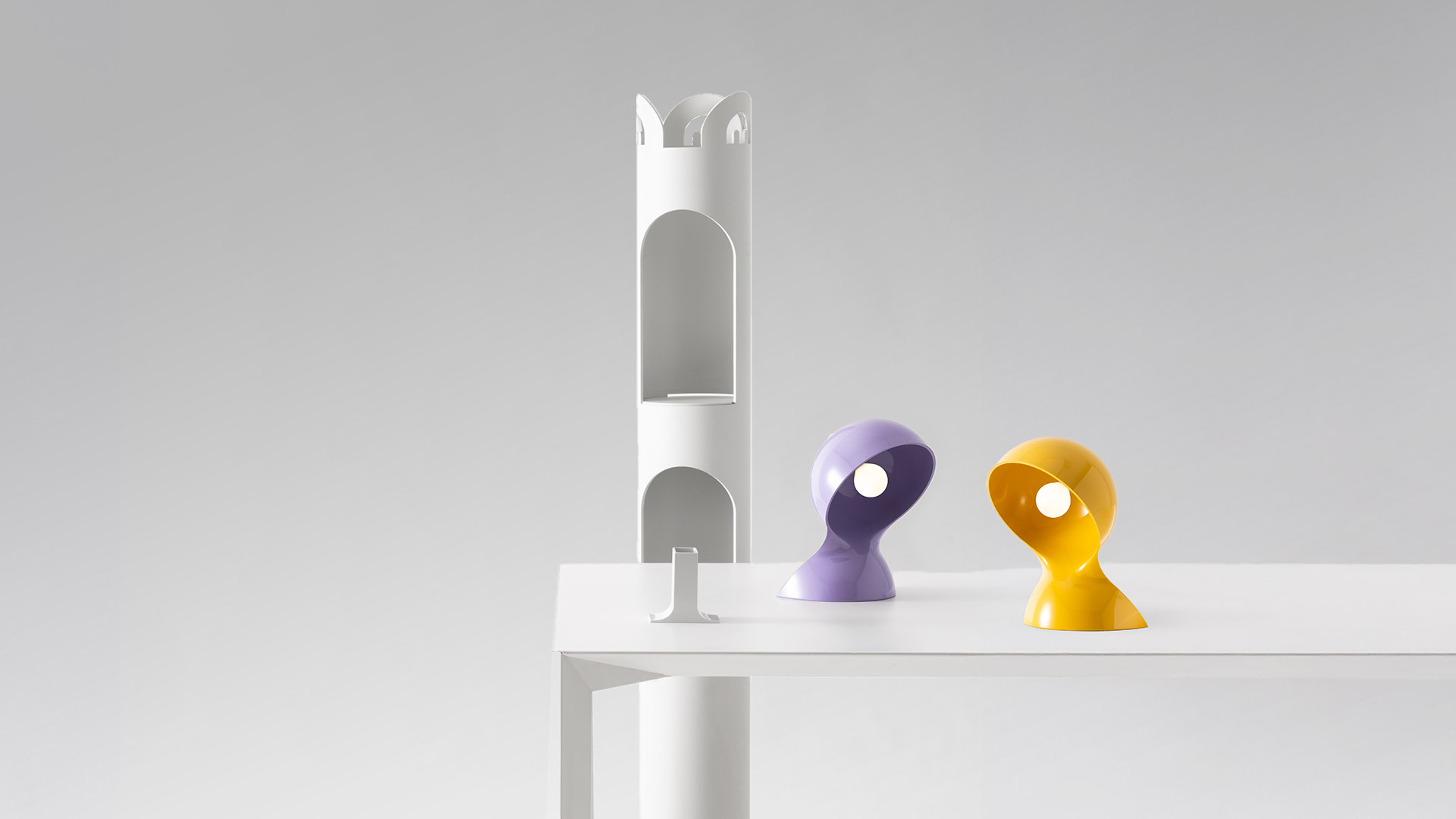 Artemide Dalù white table lamp with curved organic shape, glowing warmly on stacked books beside white flowers and a plant
