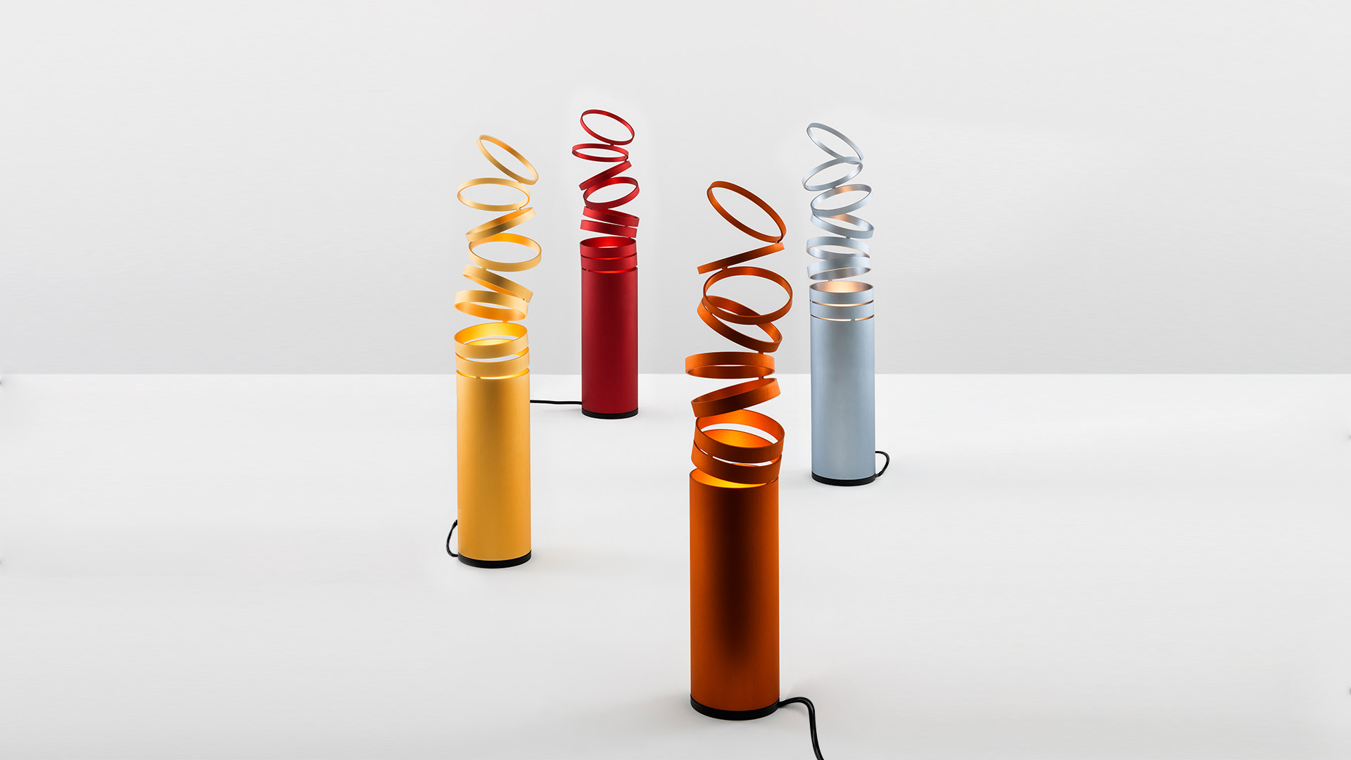 Four Artemide Decomposé Light Tavolo floor lamps with cylindrical bases and spiraling ribbon shades in yellow, red, orange, and gray