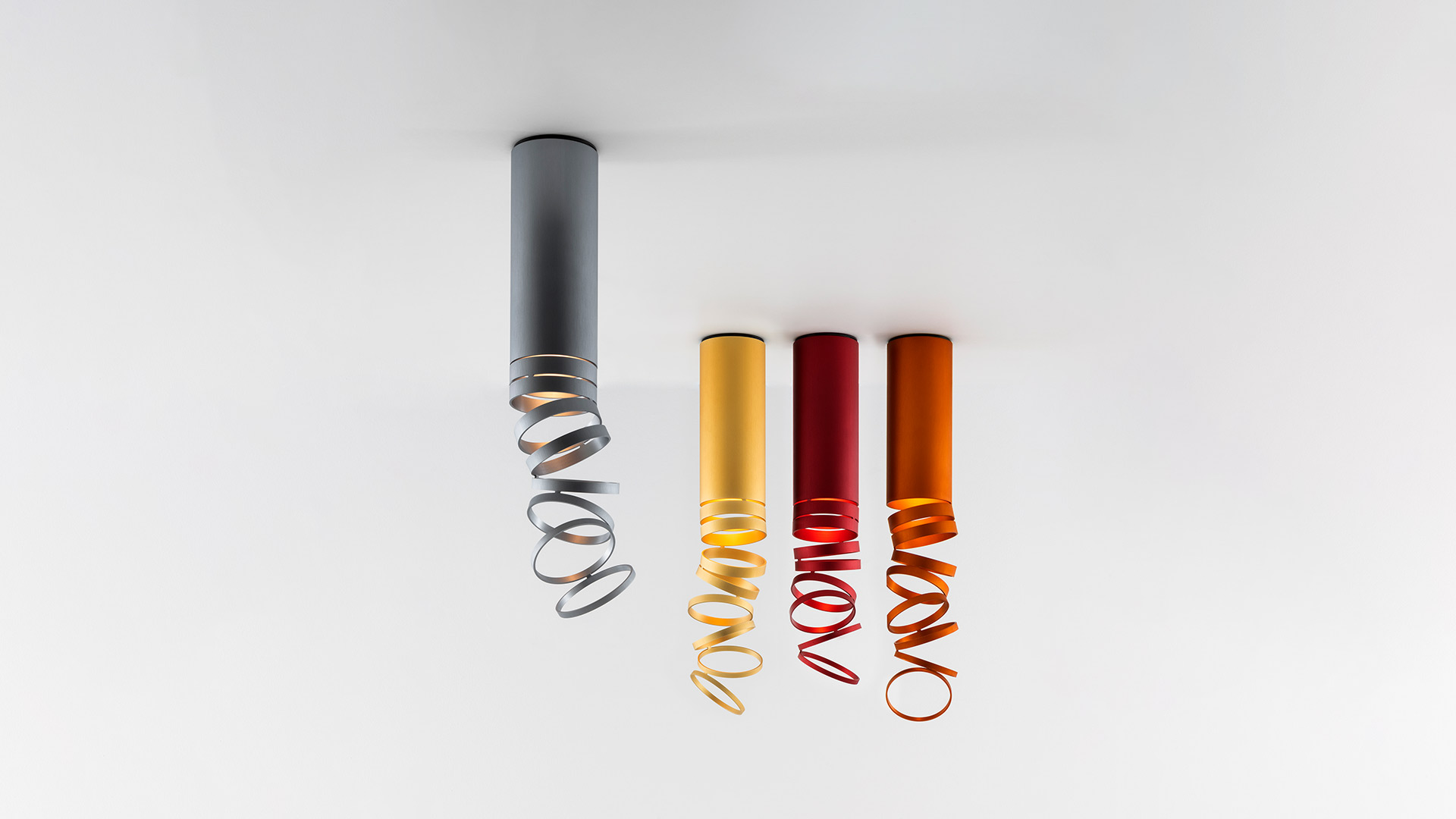 Artemide Decomposé Light Soffitto ceiling lamps in four colors: grey, yellow, red, and orange, each with a cylindrical body and spiral ribbon shade.