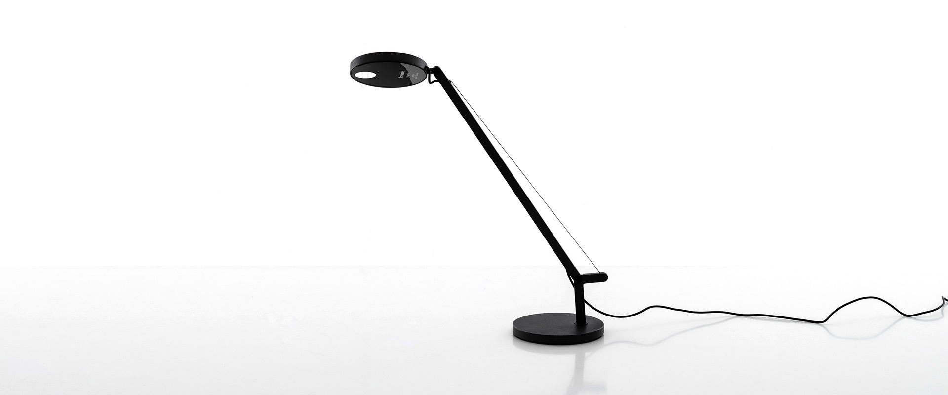 Artemide Demetra Micro table lamp in black on a wooden block, casting warm light, beside a metal shelving unit with decorative objects