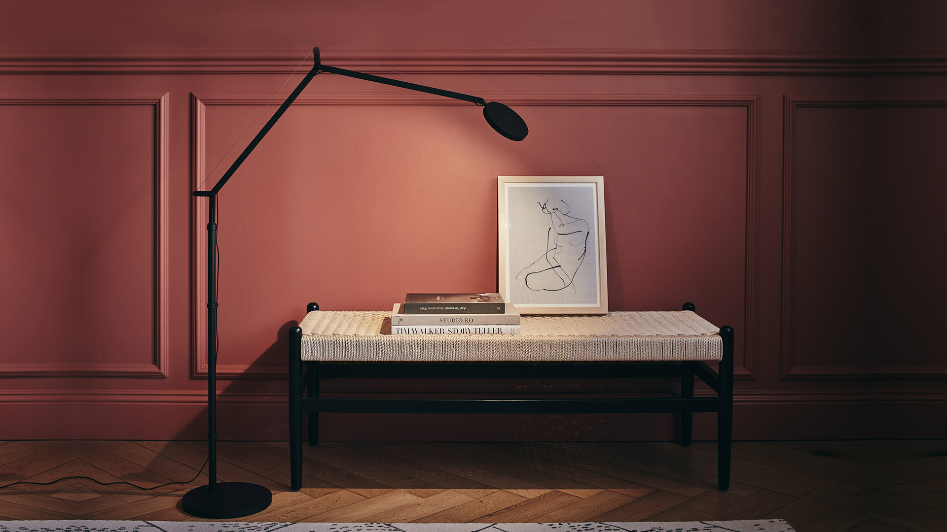 Artemide Demetra Reading Floor lamp in matte black beside a woven bench with books and framed artwork against a rose-colored wall