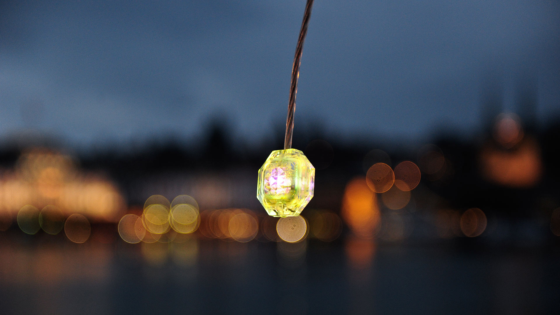 Diamante pendant light by Artemide: small faceted yellow-green gem-shaped LED lamp hanging against a blurred dusk cityscape