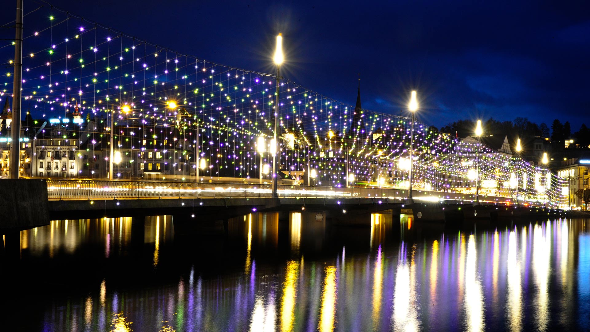 Bridge illuminated at night with multicolored Artemide Diamante string lights reflected in the river below, creating a festive urban display