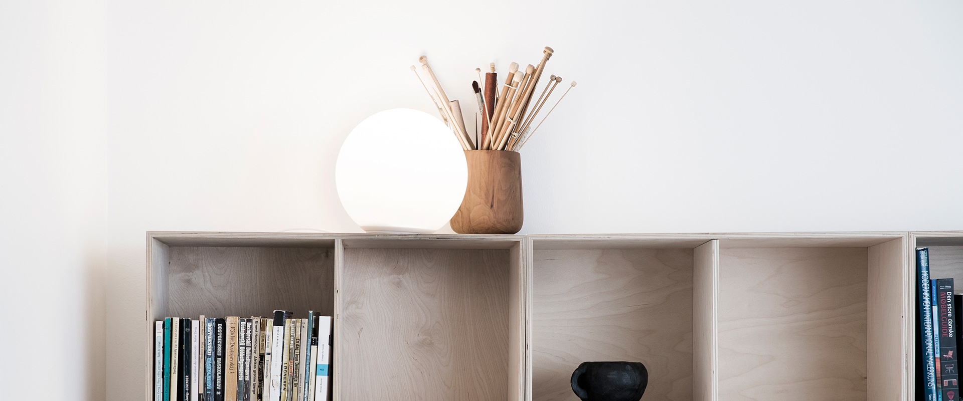 Artemide Dioscuri table lamp with opal white spherical diffuser on a wooden shelf beside a cup holding pencils and books