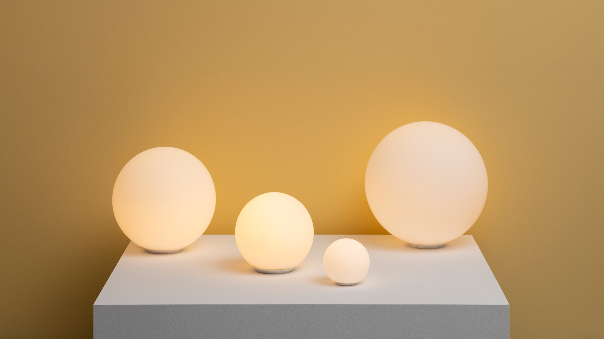 Four Artemide Dioscuri table lamps in varying sizes, opal white glass spheres glowing warmly on a wooden platform by a seaside window