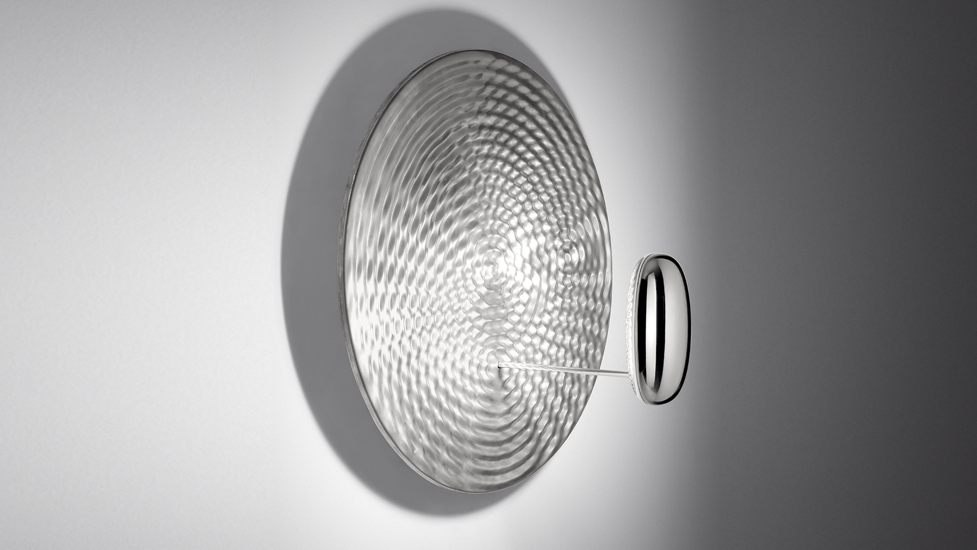 Artemide Droplet wall lamp: large circular silver disc with ripple texture mounted on wall, small oval mirror accent piece on side