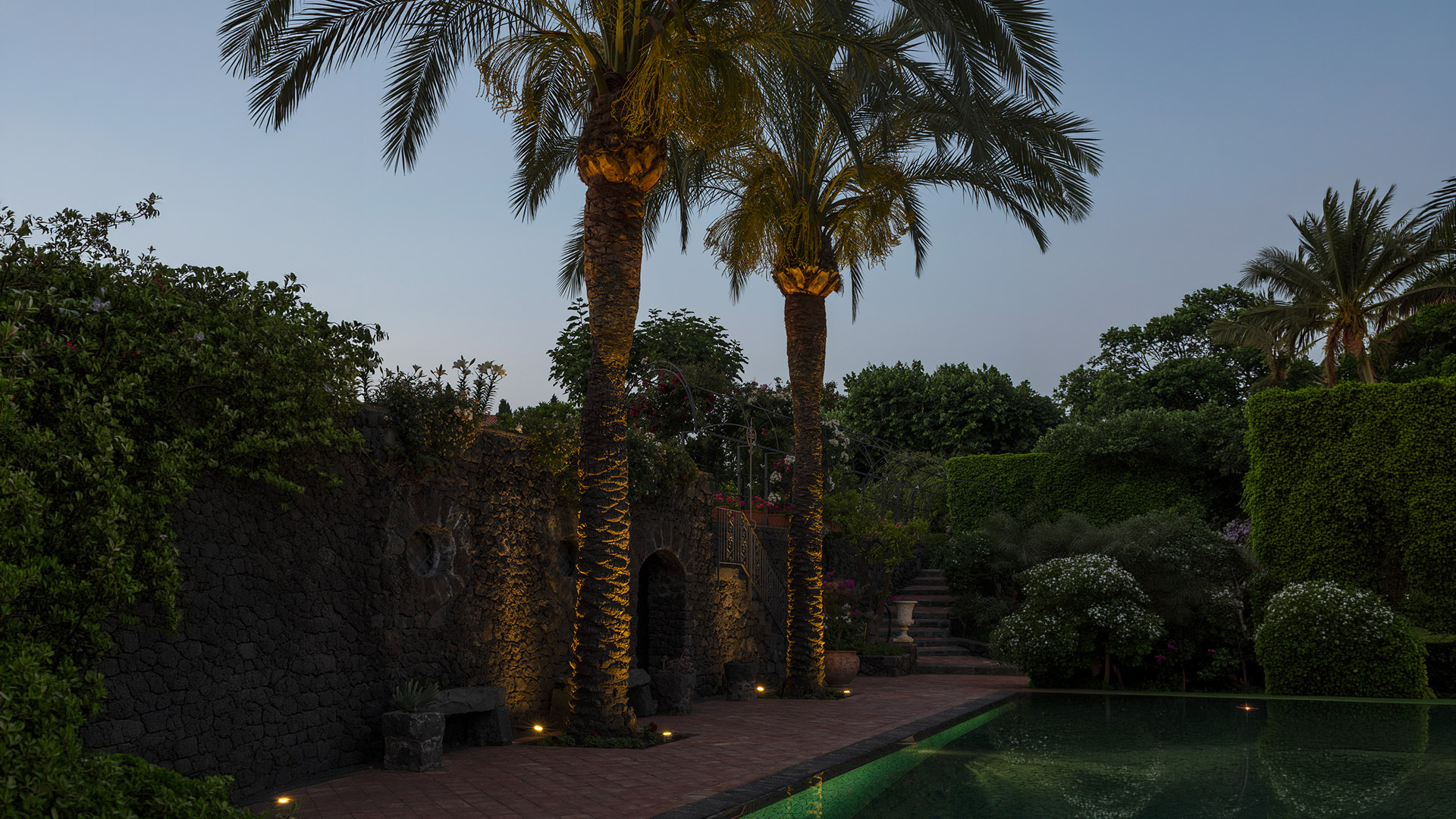 Garden at dusk with palm trees illuminated by Artemide Ego Spot ground fixtures, reflecting in a pool beside a stone wall