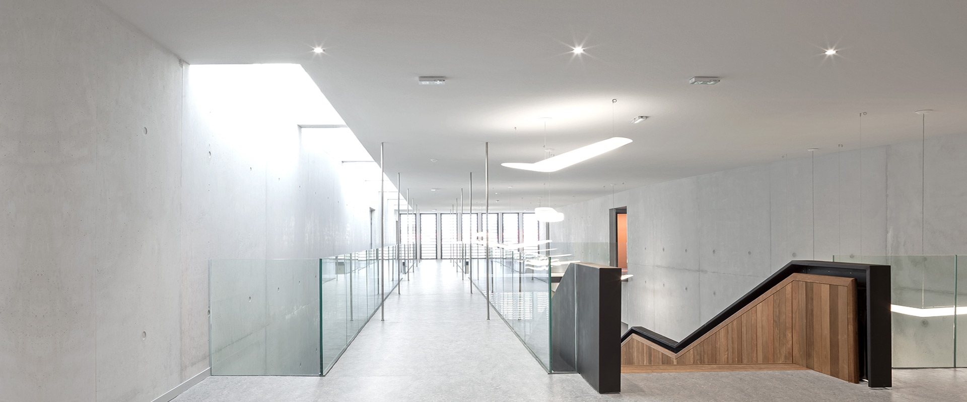 Modern corridor with Artemide Everything 55 recessed ceiling lights illuminating concrete walls, glass railings, and a wooden staircase.