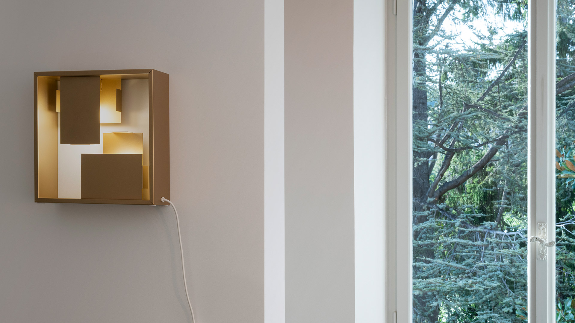 Artemide Fato table lamp in brushed brass, square frame emitting warm light, displayed on a wooden shelf with decorative sculptures