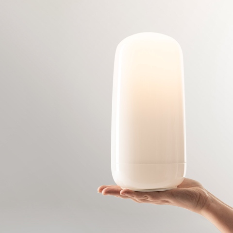 Hand holding the Artemide Gople Portable lamp, a compact white opal diffuser with soft rounded shape emitting warm light