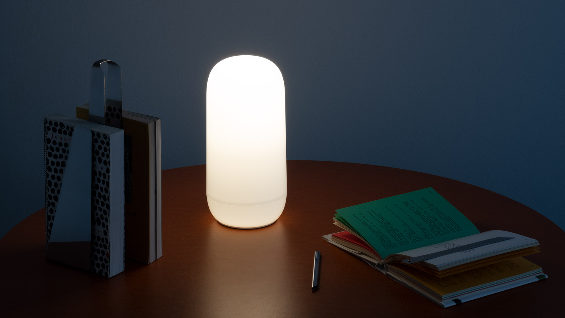 Artemide Gople Portable lamp glowing on a round wooden table, beside books and a notebook, casting warm light in a dim room