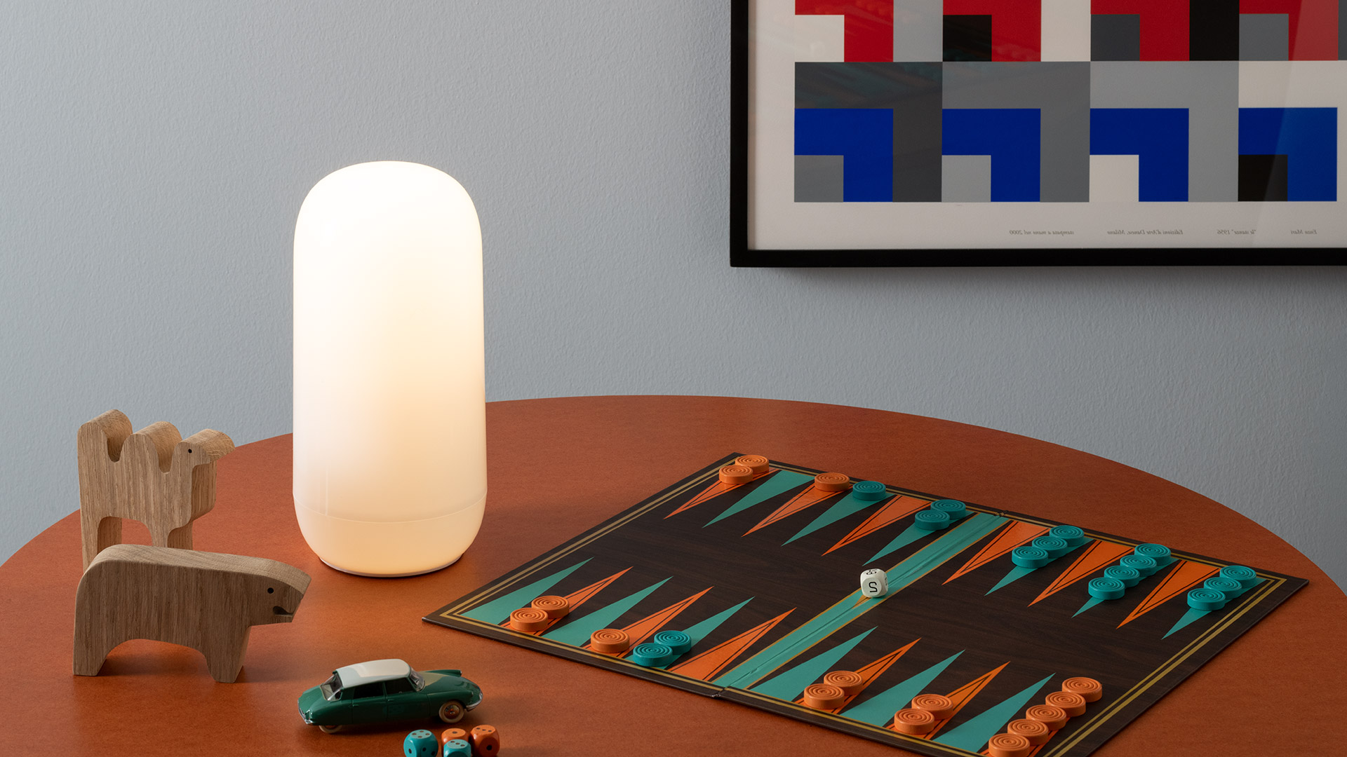 Artemide Gople Portable lamp glowing warmly outdoors at dusk, placed on grass beside an open book and a cup on a striped blanket