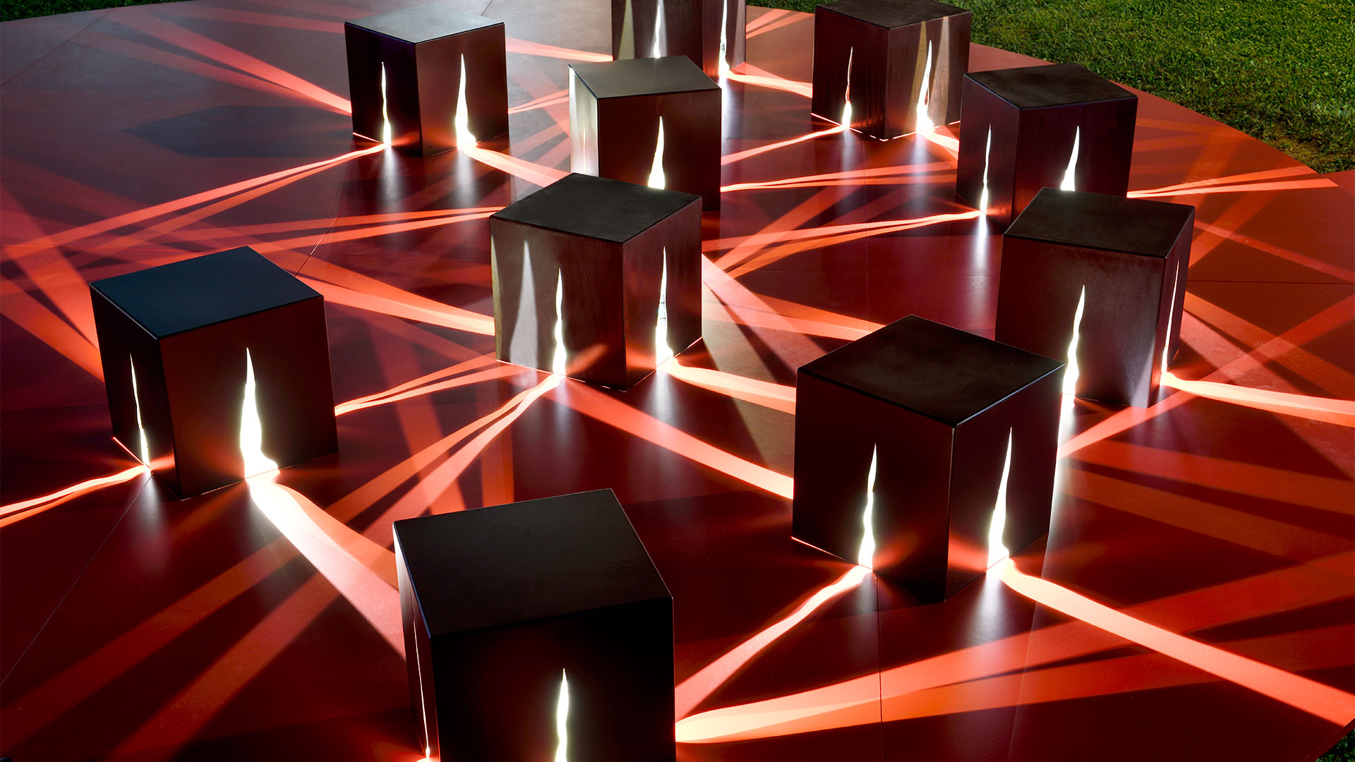 Three Artemide Granito outdoor bollard lights on grass, emitting warm light upward, with mountains and blue sky in background