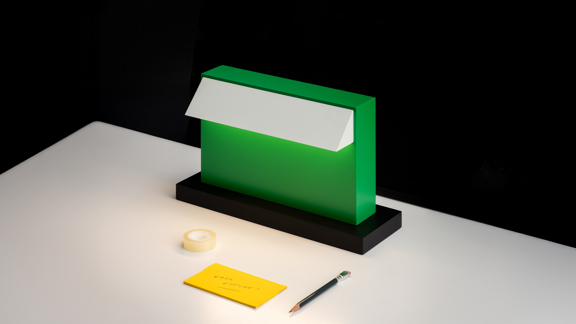 Artemide Hera table lamp: compact green rectangular body with white tilted reflector on black base, shown beside a red apple for scale