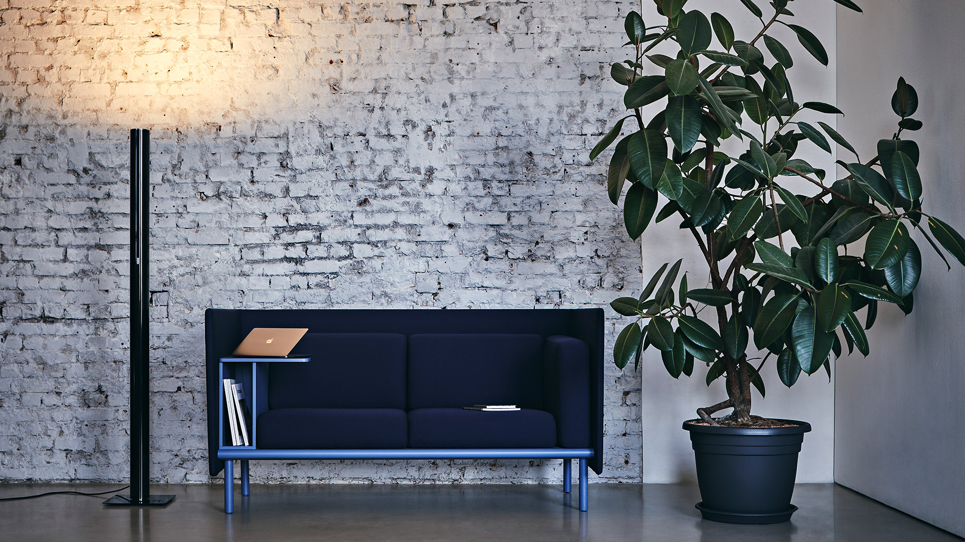 Artemide Ilio floor lamp in black beside a navy blue sofa with wooden shelf, against a white brick wall with a potted plant