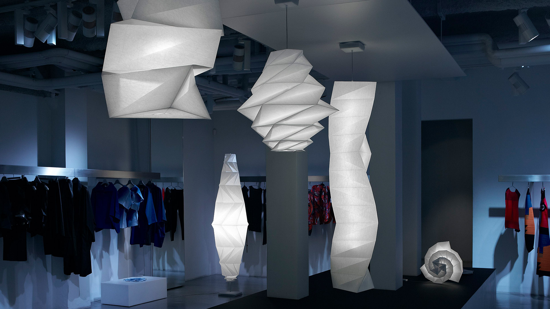 Three Artemide In-Ei Issey Miyake lamps: two origami-folded pendant lights and one spiral floor piece, glowing white against black background