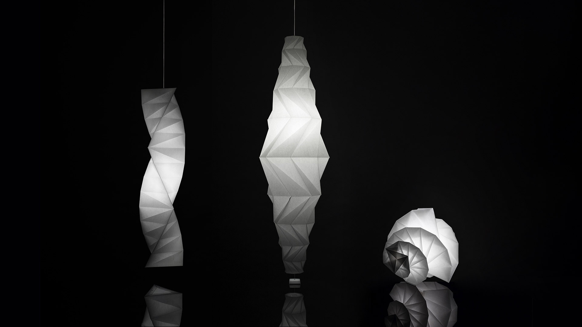Three Artemide In-Ei Issey Miyake Mendori lights: two pendant lamps and one floor lamp with origami-folded white diffusers glowing against black background