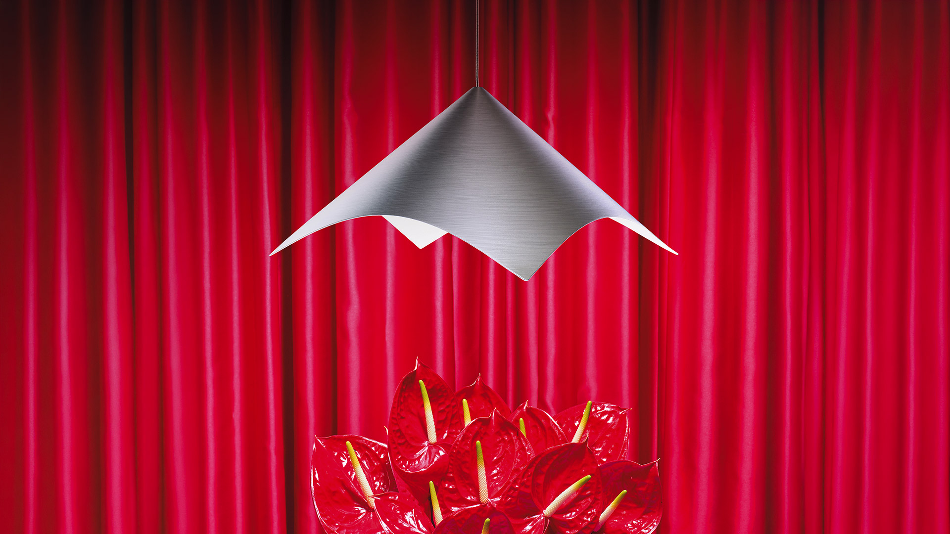 Artemide Iperbole pendant lamp with angular silver shade suspended above red anthurium flowers against a red curtain backdrop
