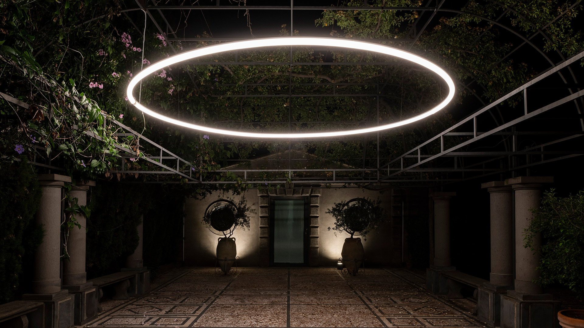 Artemide La Linea pendant lamp with sinuous glowing ribbon shape, suspended among lush green wisteria branches in an outdoor pergola