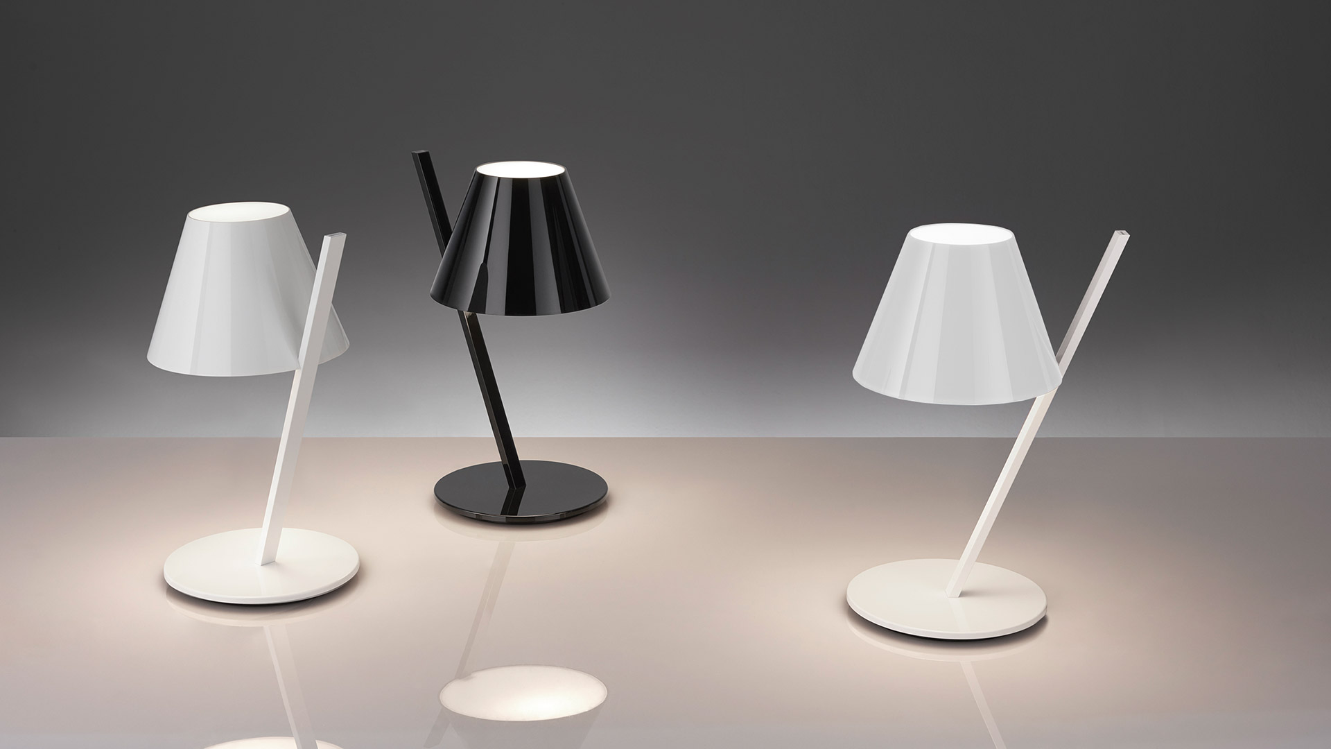 Artemide La Petite table lamp in white with conical shade on angled stem, glowing warmly on a wooden desk with books and plant