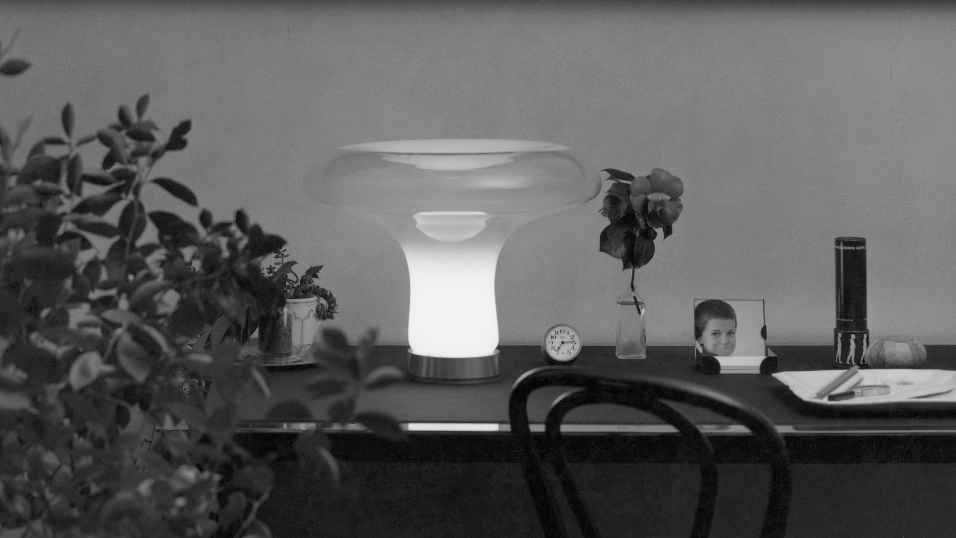 Artemide Lesbo table lamp with frosted glass mushroom-shaped diffuser and chrome base, glowing warmly on a white pedestal