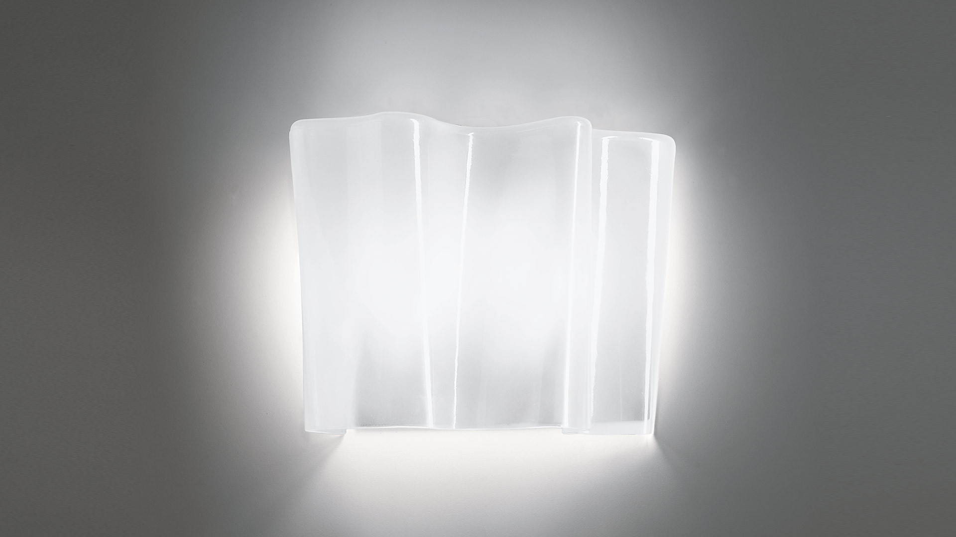 Artemide Logico wall lamp with layered frosted glass panels emitting soft diffused white light against a grey wall